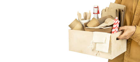 Banner with a courier holding a box with eco dishes for fast food and parasites isolated on a white background. In his hands he holds a wooden box with plates, and, cutlery, a bottle.の写真素材