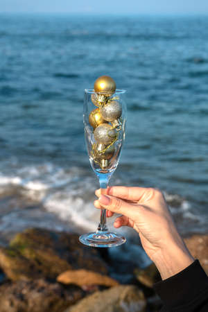 A glass with gold Christmas decorations in the hand of a young girl by the sea with a rocky shore, New Year's celebration. Vertical photoの写真素材