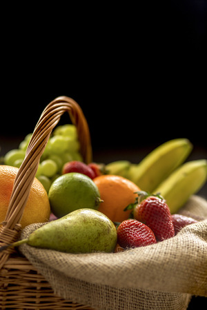 Vertical closeup detail on a basket full of fruit (banana, strawberry, pineapple, orange, pear, apple) isolated on black background - low keyの写真素材