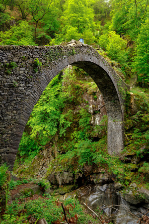 Side View on the Ponte Romano Intragna Bridge hiding in the valley with beautiful river covered with trees, close to Verzasca village. It is the oldest bridge in Switzerland.の写真素材