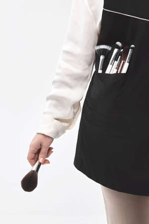 A woman professional makeup artist holds a makeup brush in her hand. The make-up artist is dressed in a working black apron in her pocket a lot of makeup brushes. Isolated on white backgroundの写真素材