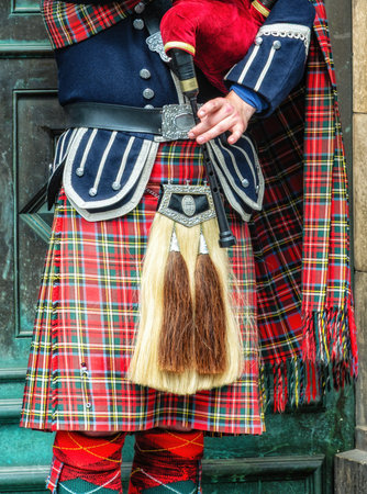 Scottish bagpiper dressed in traditional red and black tartan dress stand before stone wall. Edinburgh, the most popular tourist city destination in Scotland.の写真素材