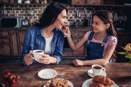 portrait of smiling girl feeding mother with cookie while drinking tea together in the kitchenの写真素材