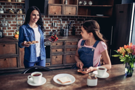 smiling daughter spreading toast with chocolate paste and looking on mother in the kitchenの写真素材