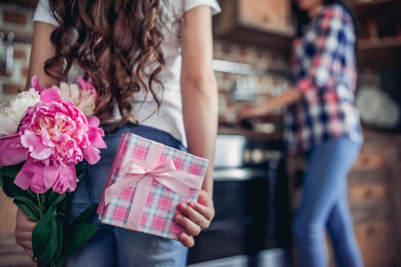 close up shot of daughter holding bouquet of flowers and gift box behind her back while her mother cooking on the stove in the kitchenの写真素材
