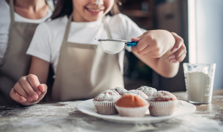cropped shot of mother and daughter sprinkling with sugar powder cooked cupcakes on the kitchen