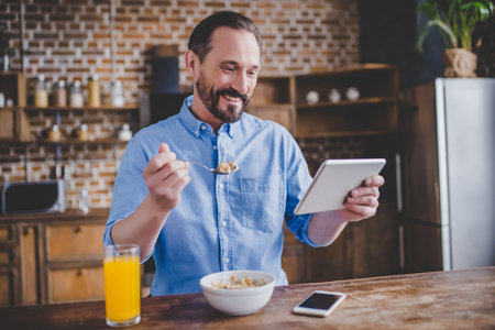 smiling bearded man eating cereals for breakfast and using digital tablet at the kitchenの写真素材