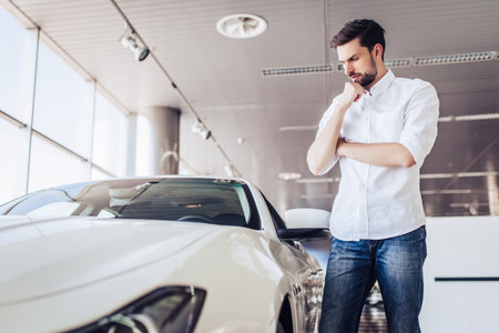 portrait of pensive male client looking at car in dealership salonの写真素材