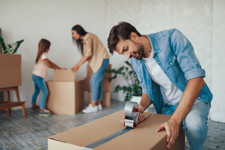 Concentrated young man putting tape on a cardboard box while whole family preparing to move out of the houseの写真素材