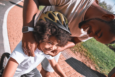 Close-up photo of a smiley Afro-American man adjusting helmet while his kid riding a bicycleの写真素材