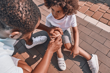 Cute Afro-American kid looking at his father putting a bandage on his knees while sitting on a pavementの写真素材