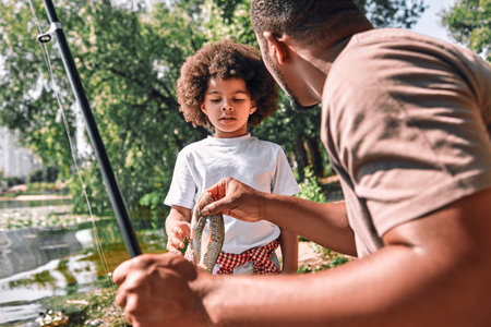 Cute Afro-American child looking st the fish in the hands of his dad on a fishing tripの写真素材
