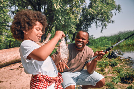 Cute curly Afro-American boy looking at the fish in his hands while his dad holding a fishing rodの写真素材