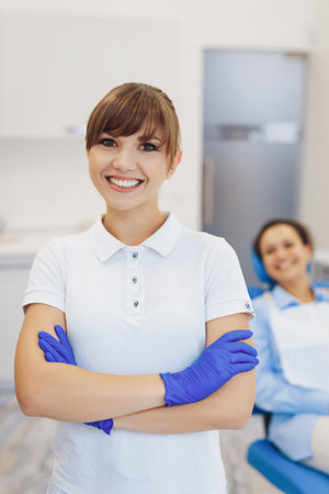 Portrait of positive dental doctor smiling at camera in dentistry room. Female doctor with crossed hands wearing latex glovesの写真素材
