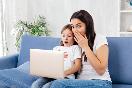 Charming woman and lovely daughter expressing surprise while watching a film on laptopの写真素材