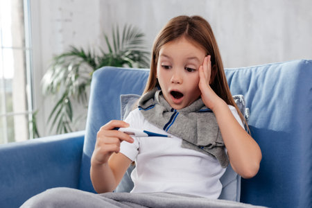 Astonished child sitting on couch with open mouth while looking at thermometer and keeping hand on...