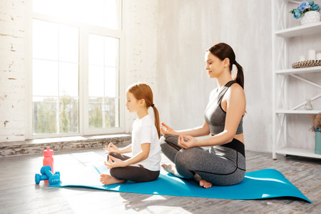 Calm girl keeping legs crossed while resting on mat and practicing yoga with her sportive mother behindの写真素材