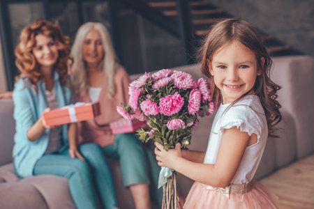 little girl holding bouquet of flowers and smiling at the camera while mother and grandmother sitting on the sofaの写真素材