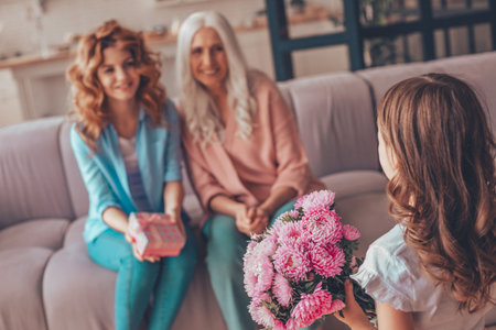 senior woman and adult child sitting on the couch while little girl congratulating them with bouquet of flowersの写真素材