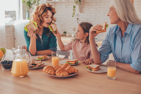 girl feeding mother with sandwich while granny sitting near and having breakfastの写真素材