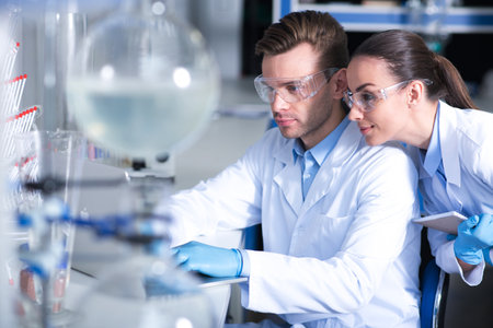 computerized researches. Two young pleasant responsible scientists in a glasses and rubber gloves working in the laboratory with the laptop looking in the screen and writing results of the research.の写真素材