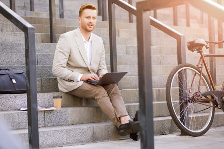 Charismatic man in classic suit working with the laptop and being thoughtful while sitting on stairsの写真素材