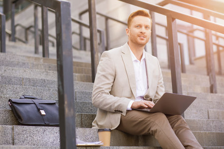 Optimistic businessman looking away and holding a laptop on knees while sending messages to his clientsの写真素材