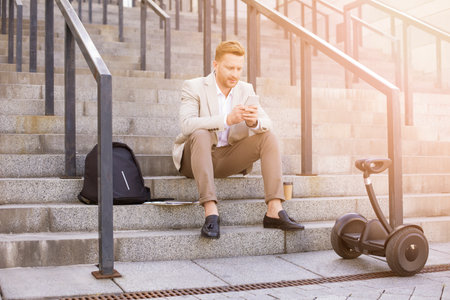 Handsome businessman using a smartphone and sitting on outdoor stairs while reading messages