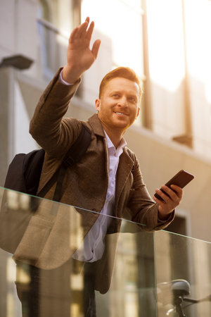 Excited businessman greeting his friend with a hand gesture and holding mobile phone in handの写真素材