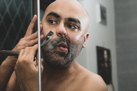 A bald, British Asian man, grooming and sculpting his beard at home in the bathroom, in front of the mirror. He is using a traditional cut throat razor or a barber's razor. This can be used to theme issues around maleness, dating and middle age.の写真素材