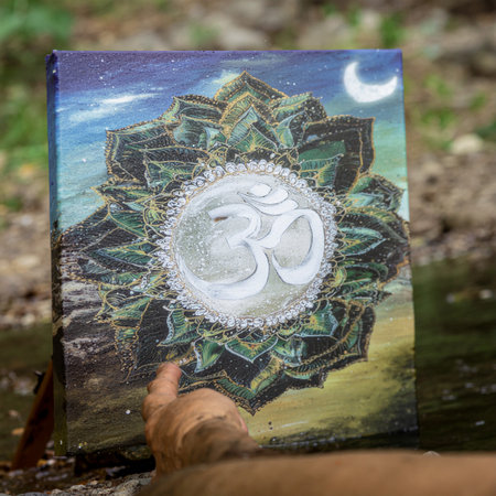 A handmade painting of a spiritual Om symbol on a mandala canvas, placed outdoors by a river. A beautiful concept of art harmonizing with nature's elementsの素材
