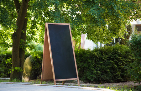 A blank blackboard sign standing on the streetside, with plants and trees around.の写真素材