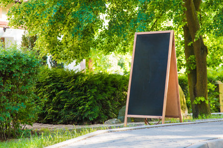A blank blackboard sign standing on the streetside, with plants and trees around.の写真素材