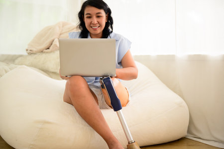 Asian young woman using laptop and relaxing at home, Positive woman with prosthetic leg having fun on weekend at home, Leisure lifestyles and activities at home, Woman working at home officeの写真素材