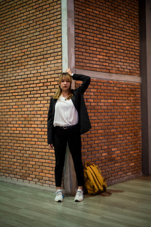 Blond hair woman in black and white clothes standing at the corner of brick wall with yellow backpack on the ground, moody and upset.の写真素材