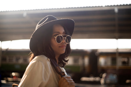 Wavy hair woman in white shirt and black hat hang backpack on her shoulder at train platform with sunlight waiting for the train to the destination.の写真素材
