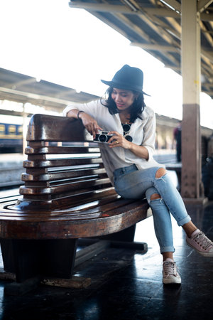 Wavy hair woman in black hat sitting on the bench holding the film camera to check at train station before travel.の写真素材