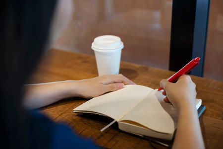 A woman sitting in the cafe with hot coffee, open the notebook and writing her diary while waiting for her lover as an appointment, over shoulder shot.の写真素材