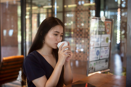 Long hair woman in dark blue dress sitting in the cafe waiting for her boyfriend, drinking hot coffee, hold a cup of coffee tightly.の写真素材