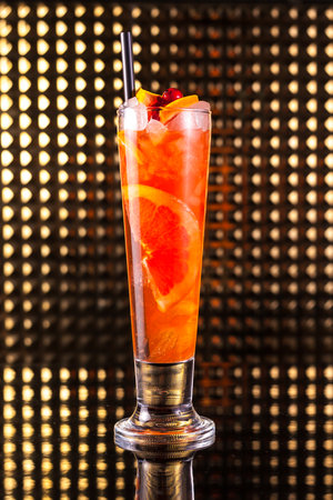 Red fruit cocktail with orange in tall glass with ice cubes on golden backdropの写真素材