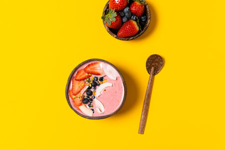 Spring diet, body detox, healthy breakfast concept, lifestyle blog - coconut bowl with fruit smoothie, fresh strawberries, blueberries, coconut chips, top view, yellow backgroundの写真素材