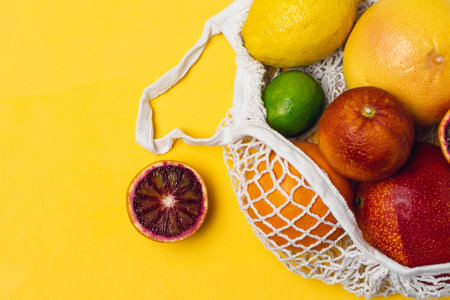 Organic citrus fruits variety in cotton mesh reusable shopping bag, bloody orange, lemon, lime, grapefruit, yellow background, top view - recycling, sustainable lifestyle, zero waste, no plasticの写真素材