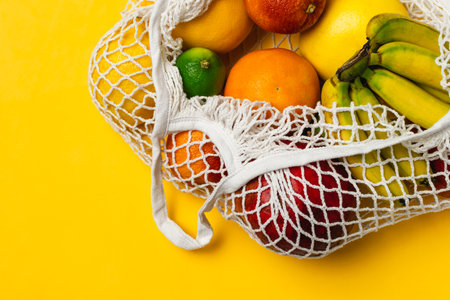 Organic fruits variety in cotton mesh reusable shopping bag, banana, mango, orange, lime, grapefruit, yellow background, top view, copy space - recycling, sustainable lifestyle, zero waste, no plasticの写真素材