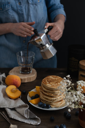 Female hands pouring coffee from moka coffe machine into a glass, Beautiful served rustic breakfast - stack of american pancakes with blueberries, peach, Gypsophila flowers, , wooden tableの写真素材