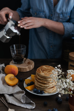Female hands pouring coffee from moka coffe machine into a glass, Beautiful served rustic breakfast - stack of american pancakes with blueberries, peach, Gypsophila flowers, , wooden tableの写真素材