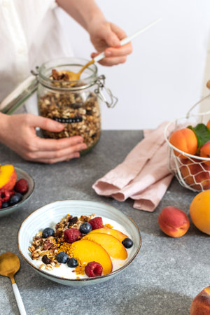 Female hands serving granola from jar, granola with greek yogurt fresh peach, blueberry, raspberry on the table - healthy breakfast conceptの写真素材