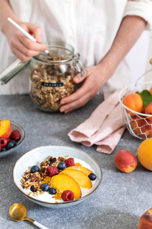 Female hands serving granola from jar, granola with greek yogurt fresh peach, blueberry, raspberry on the table - healthy breakfast conceptの写真素材