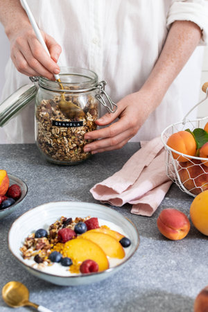 Female hands serving granola from jar, granola with greek yogurt fresh peach, blueberry, raspberry on the table - healthy breakfast conceptの写真素材