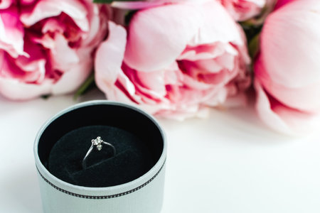 Engagement concept - diamond ring in a box, pink peonies flowers on white background, copy space
