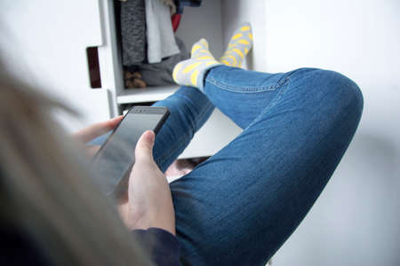 Teenage girl, using a smartphone in blue jeans and colored socks, sitting with her legs raised and resting them on the wardrobe Rear viewの写真素材